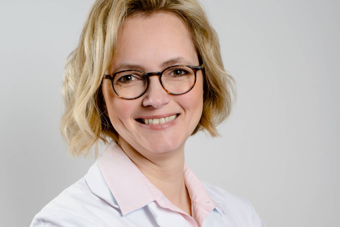 Eine lächelnde Frau mit kurzen blonden Haaren und Brille trägt einen weißen Laborkittel über einem hellrosa Hemd mit Kragen. Sie steht vor einem schlichten, hellen Hintergrund.